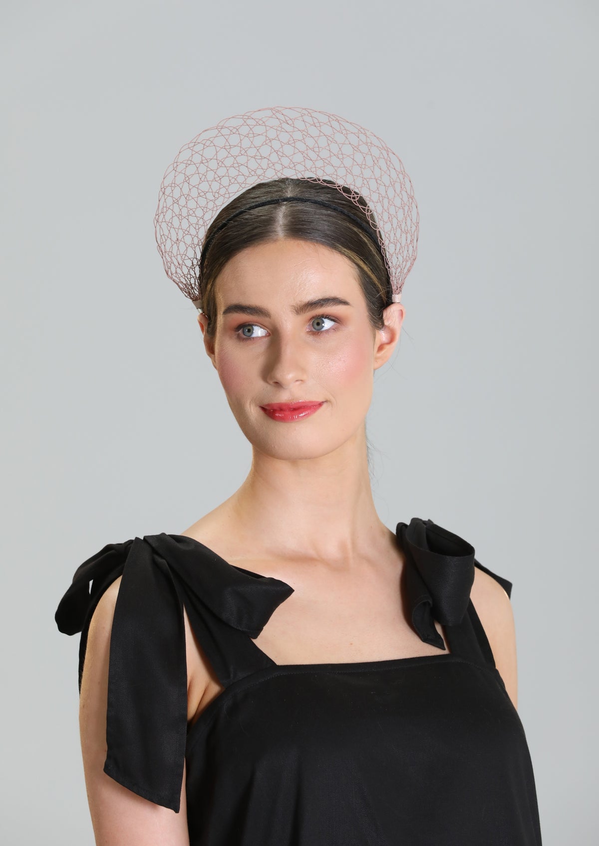 Woman wearing a black dress with bow details and a decorative dusky pink crown wire lace headpiece against a gray background