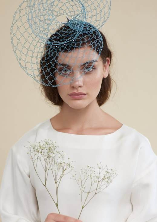A model wearing a white blouse and holding baby's breath flowers wears an asymmetric sky blue headpiece. It is wire made into lace, and is worn on the model's right hand side. It goes over her eyes and extends above her head in a rounded shape.  The lace is constructed like a wide mesh so you can see through it.