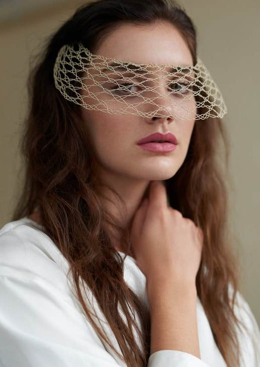 A model with long hair looks through a sand coloured wire lace veil in the shape of a visor.