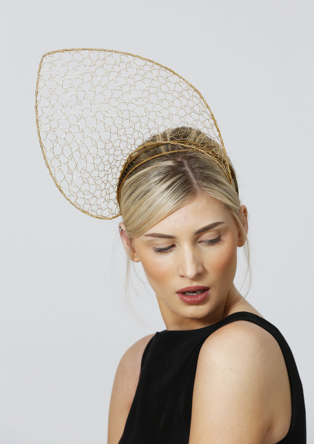 A model wearing a gold headpiece looks down over her shoulder. The headpiece is shaped like a petal and sits above her head, with a height about the same again as her head. It tilts to her right hand side. It is constructed of an outer frame in gold giving the petal outline, filled in with a finer mesh-llike gold wire lace that is see-through.
