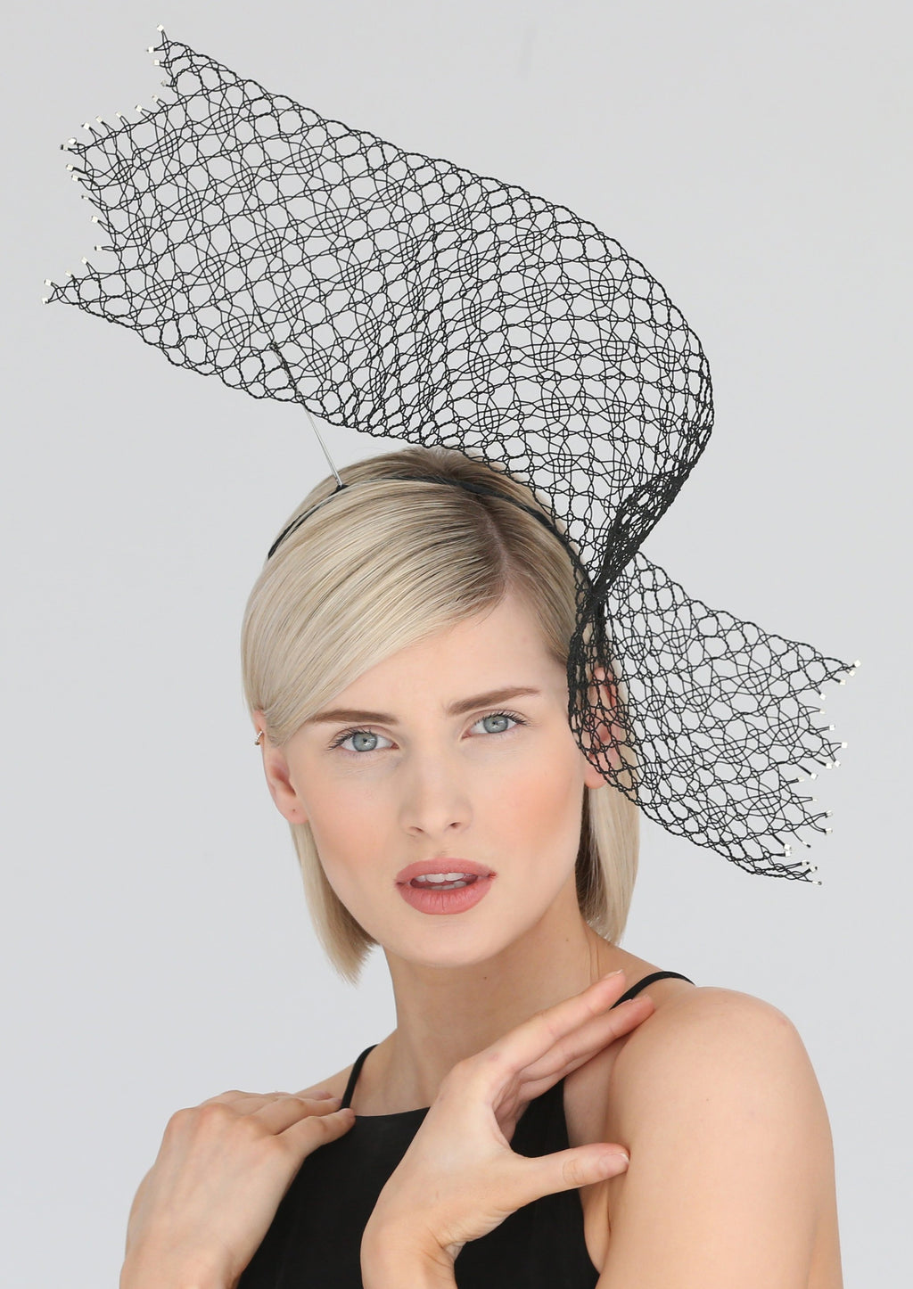 A model with blonde hair wears a black headpiece on her left hand side. It is formed of a wide rectangular piece of mesh-like wire lace in a pattern reminiscent of stars. The mesh is see-through. The ends are uneven like a zigzag and the wire ends are finished with tiny silver metal plates. The rectangle starts high above her head, curving back where it meets her head above her left ear and then curling outwards again to the side of her face.