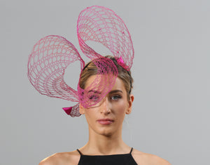 A model wears a wire lace headpiece that sits mostly to her right hand side. It loops twice above her head curling down over her right eye in between and is finished with pink satin triangles at each end. The wire is bright pink and changes tone to orange in some places in an ombré effect.