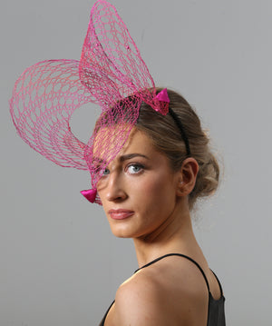 A model is shown from a side view looking over her left shoulder. She wears a wire lace headpiece positioned mostly on her right side that loops twice above the head, curling down over her right eye in between the loops, and is finished with pink satin triangles at each end. The wire is bright pink and changes tone to orange in some places in an ombré effect.