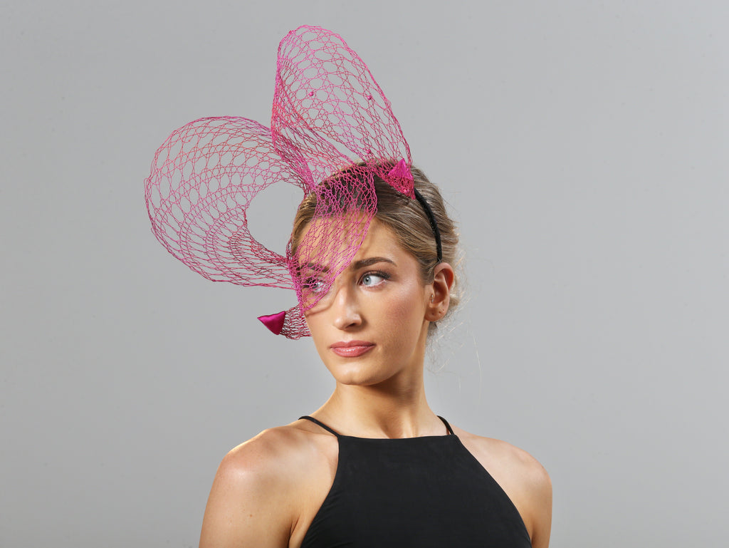 A model tilts head to her right. She wears a wire lace headpiece that is placed mostly on her right hand side. It loops twice above her head, curling down over her right eye in between loops, and is finished with pink satin triangles at each end. The wire is bright pink and changes tone to orange in some places in an ombré effect.