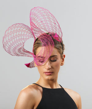 A model is looking downwards. She wears a wire lace headpiece positioned mainly to her right hand side. It loops twice above her head, curling down over her right eye in between the loops, and is finished with pink satin triangles at each end. The wire is bright pink and changes tone to orange in some places in an ombré effect.