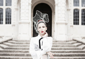 A model wears a headpiece of wired ivory veiling that sits above her head and comes down in front of her right eye. In the background are the steps leading up to an old building and the model is framed in front of the arched entrance.